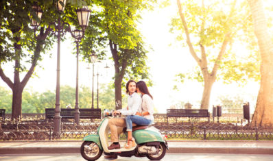 Sideview of a trendy couple riding a scooter in the morning. Sun is shining through trees Sideview of a trendy couple riding a scooter in the morning. Sun is shining through trees